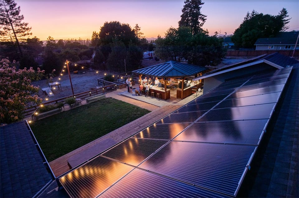 Solar panels reflecting outdoor string lights at dusk with group of people under pergola below
